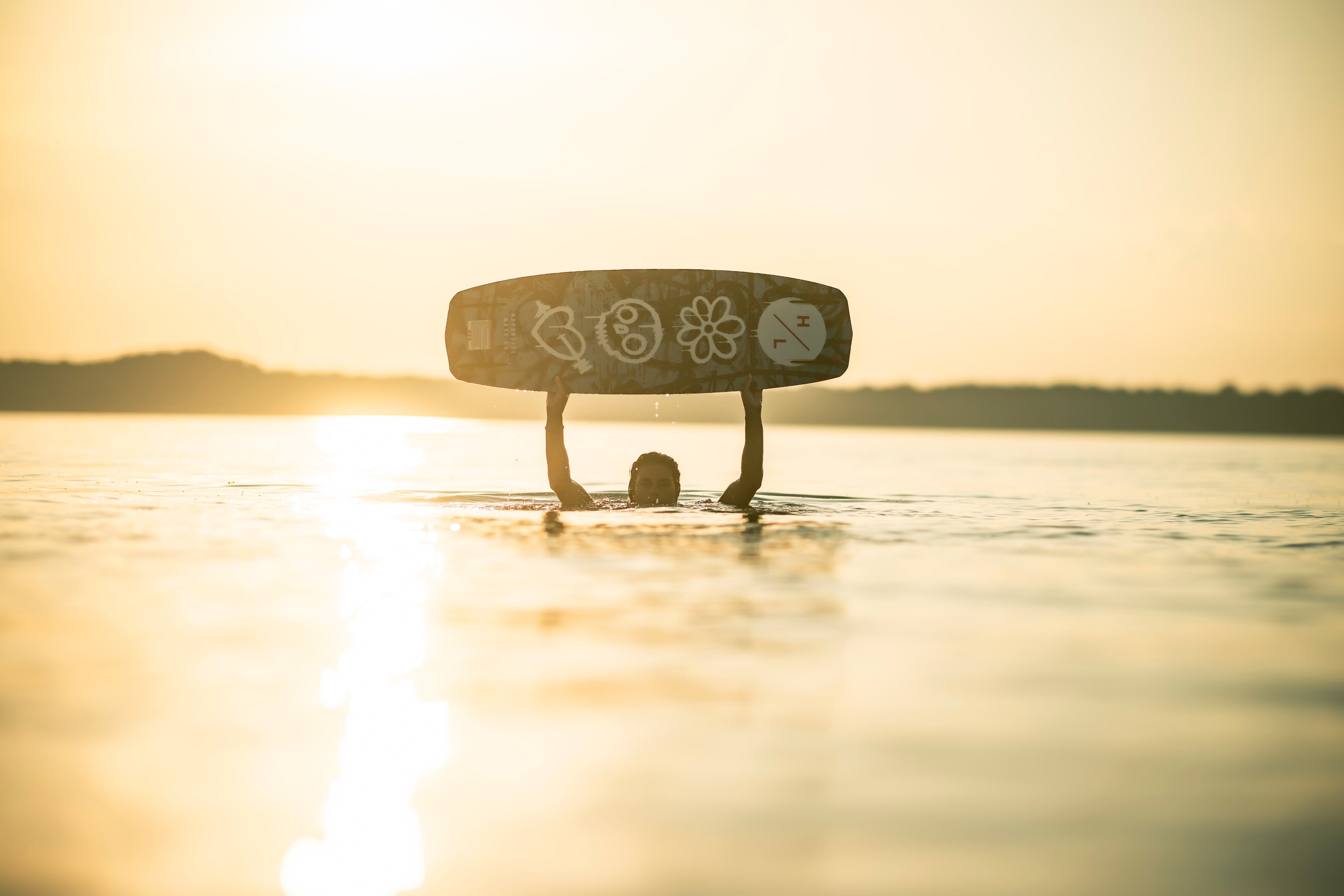 Person holding a wakeboard above their head in a sunlit lake.