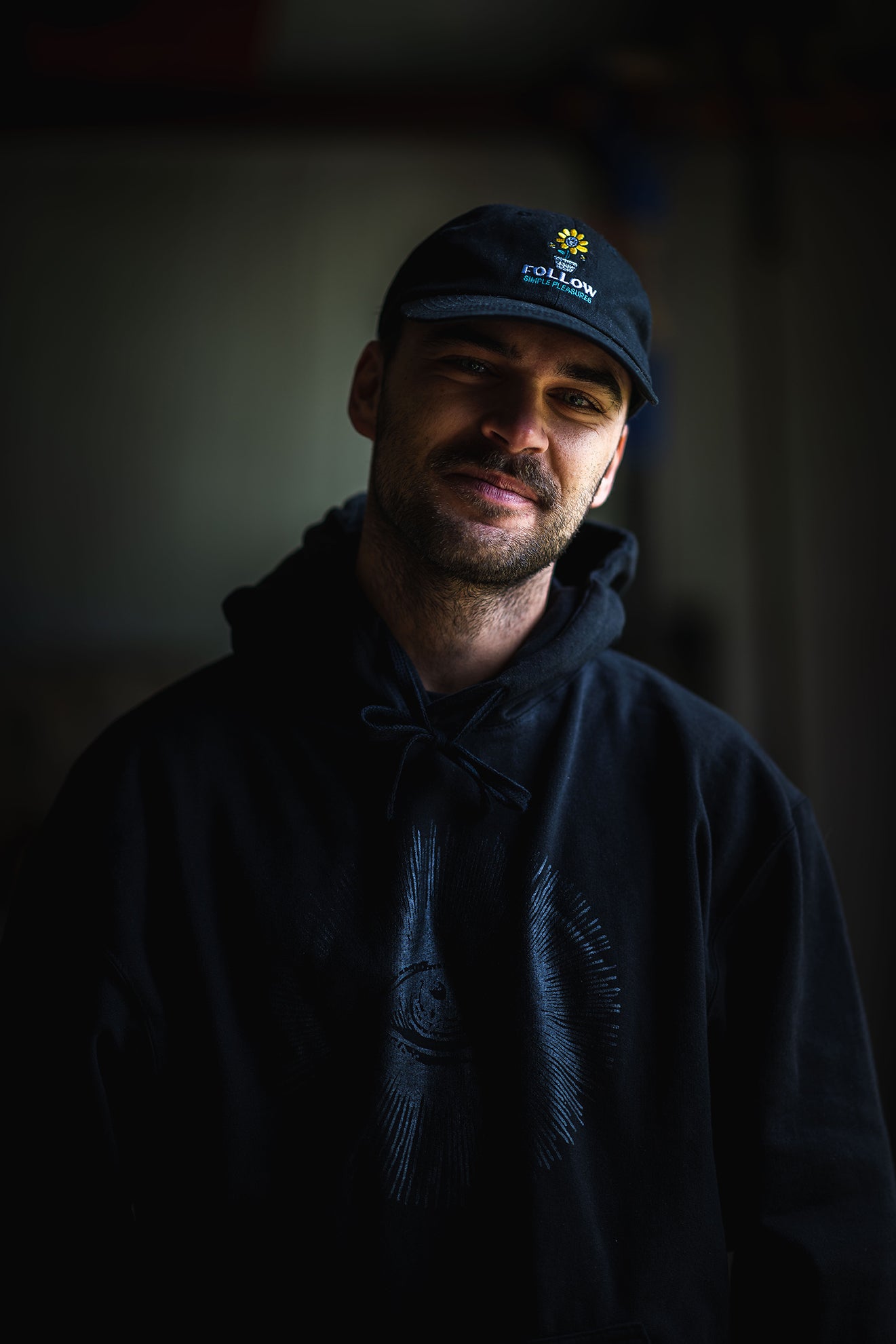 Person in a dark hoodie and cap stands in low light against a dim background.