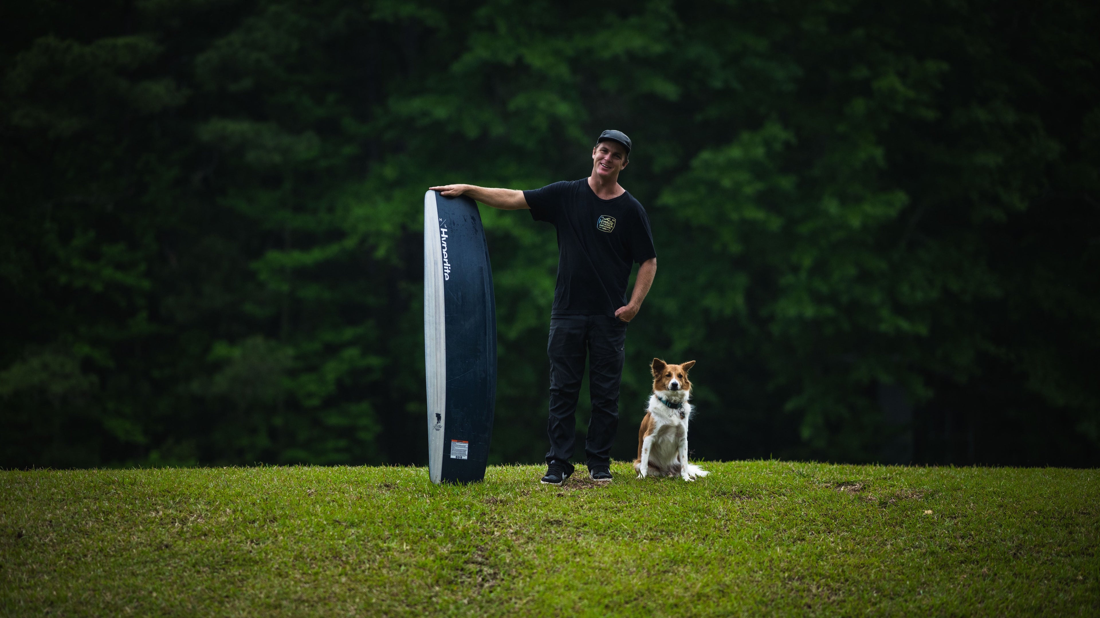 Man with surfboard and dog on grass, trees in background.