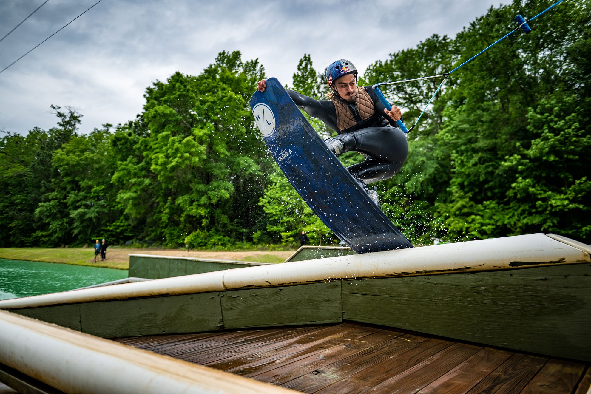 Person wakeboarding in a wetsuit, performing a jump near a wooded area.