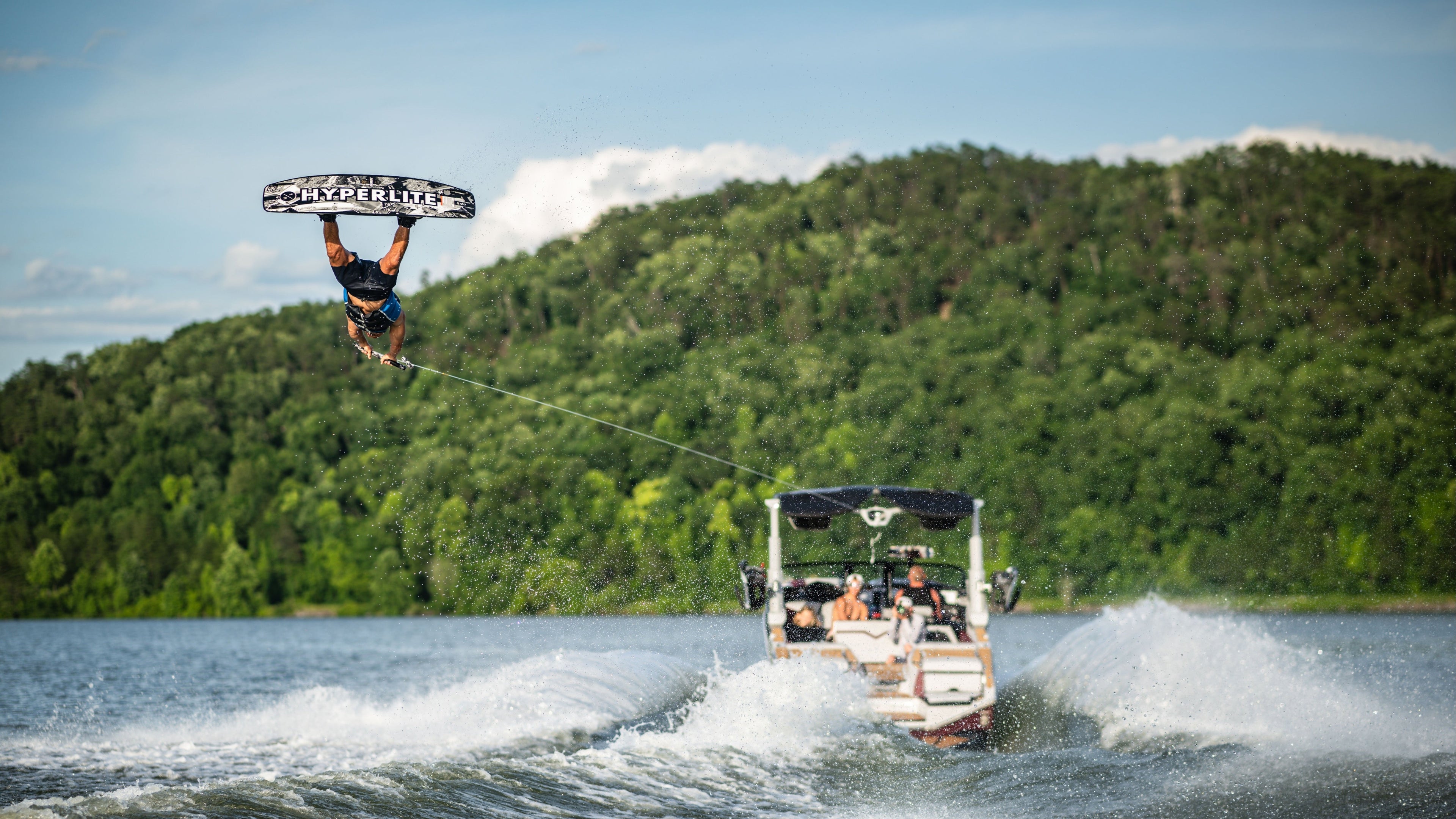 Person wakeboarding in the air, pulled by a boat on a lake with a forested background.