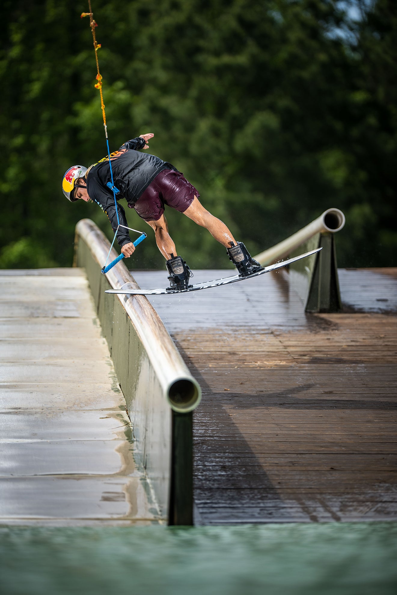 Person wakeboarding on a rail, holding a rope handle, against a forest backdrop.