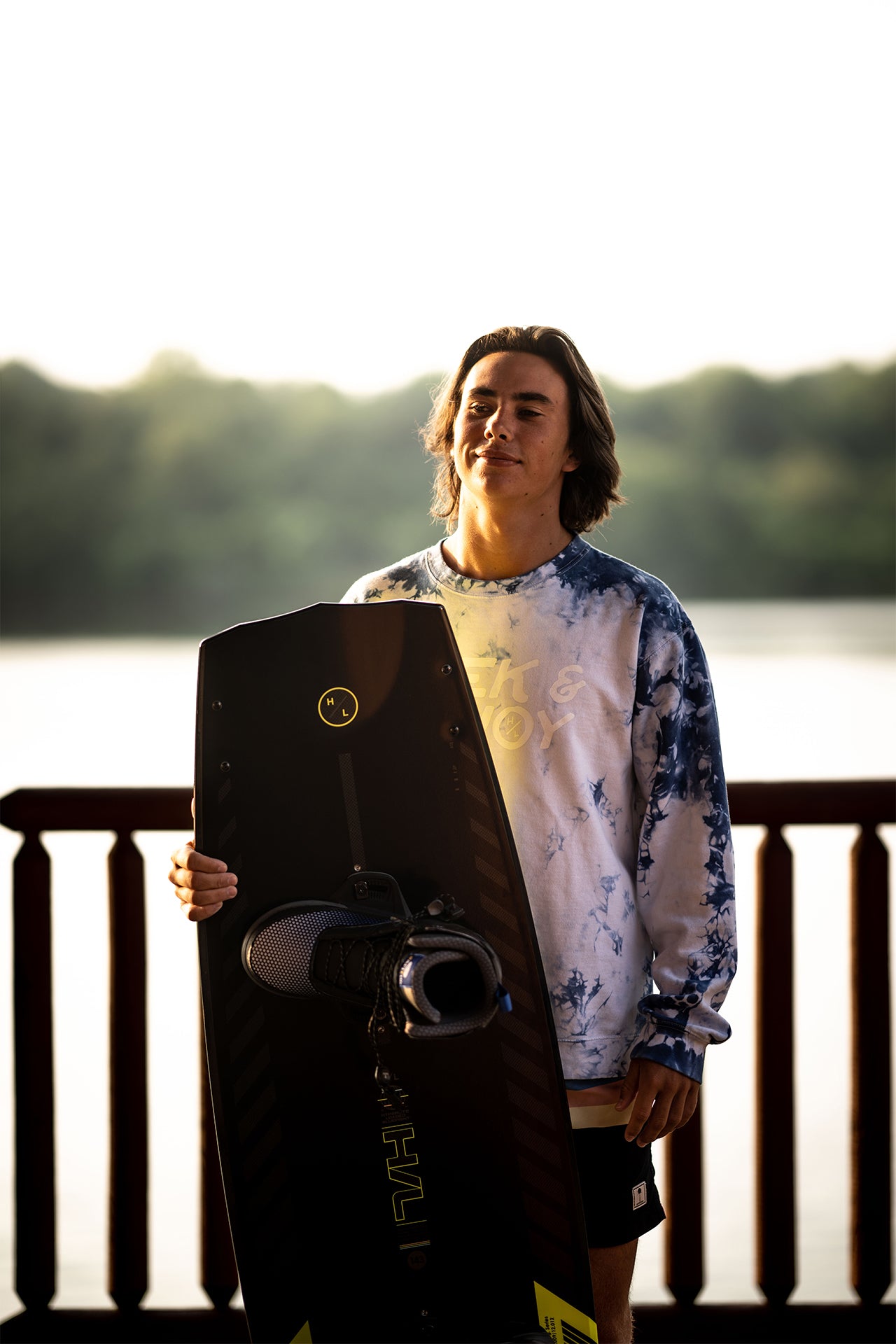 Person holding a wakeboard on a deck by the water, with trees in the background.