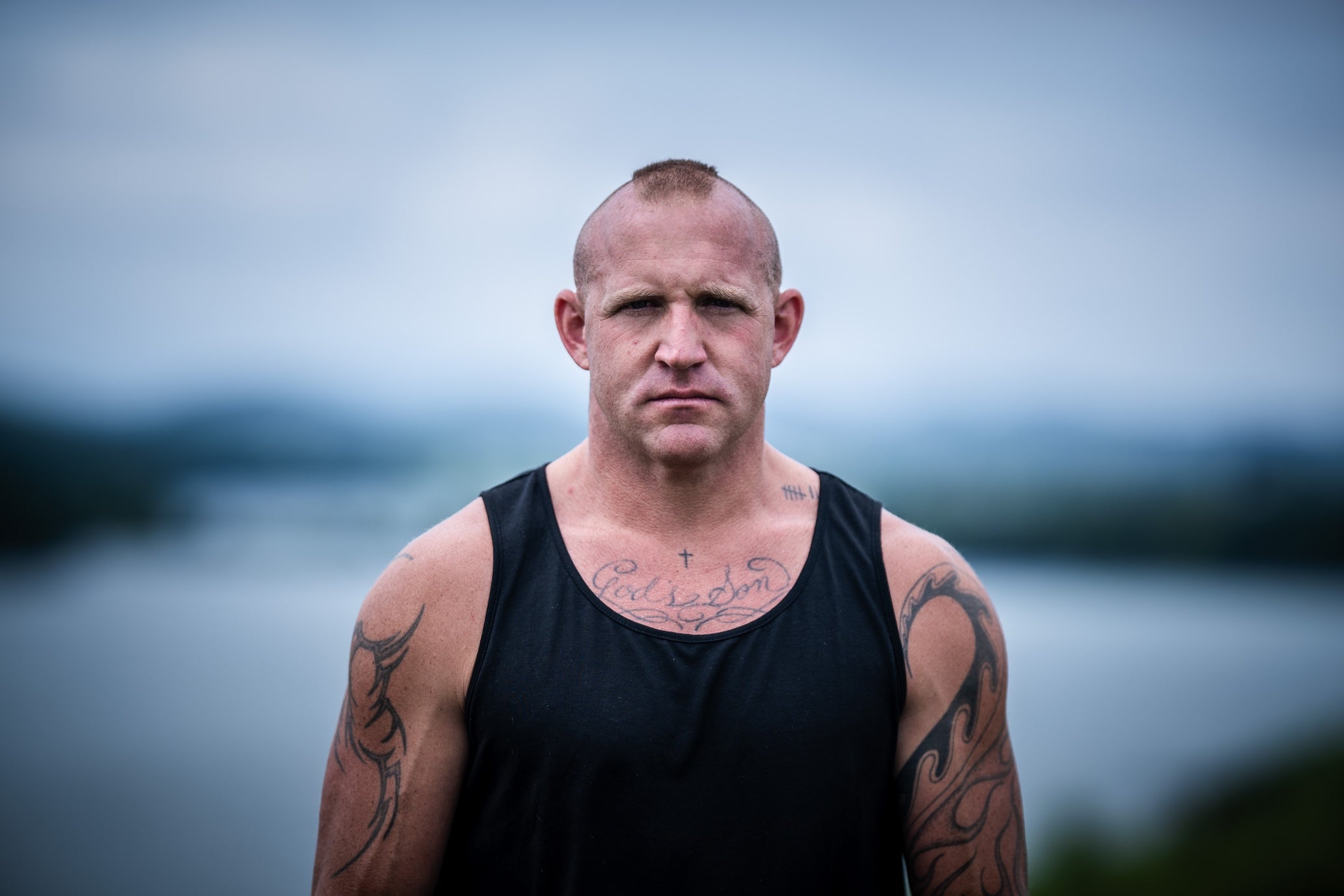 Man with tattoos wearing a black tank top against a blurred natural background.