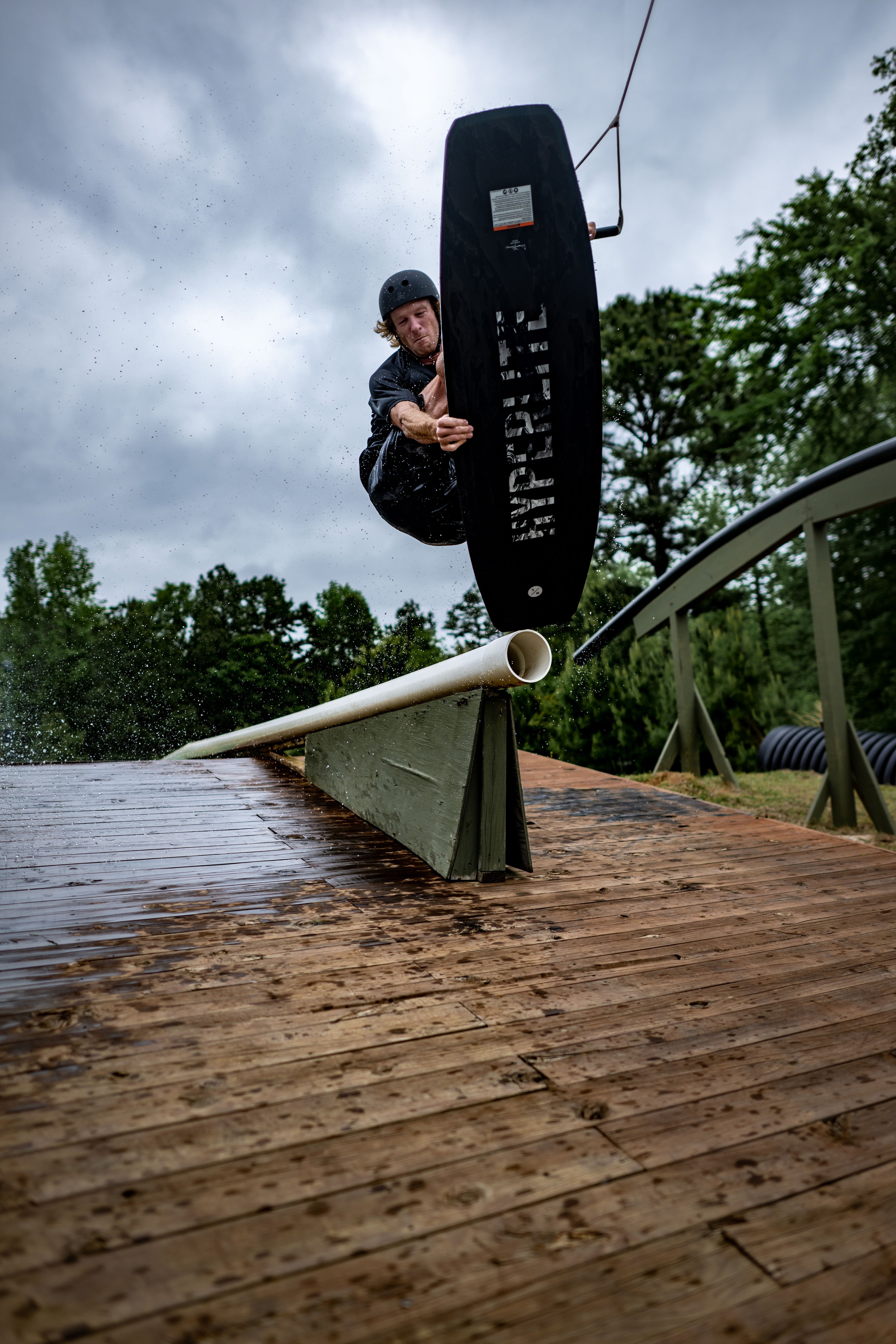 Person wakeboarding on a rail in a wooded area under a cloudy sky.