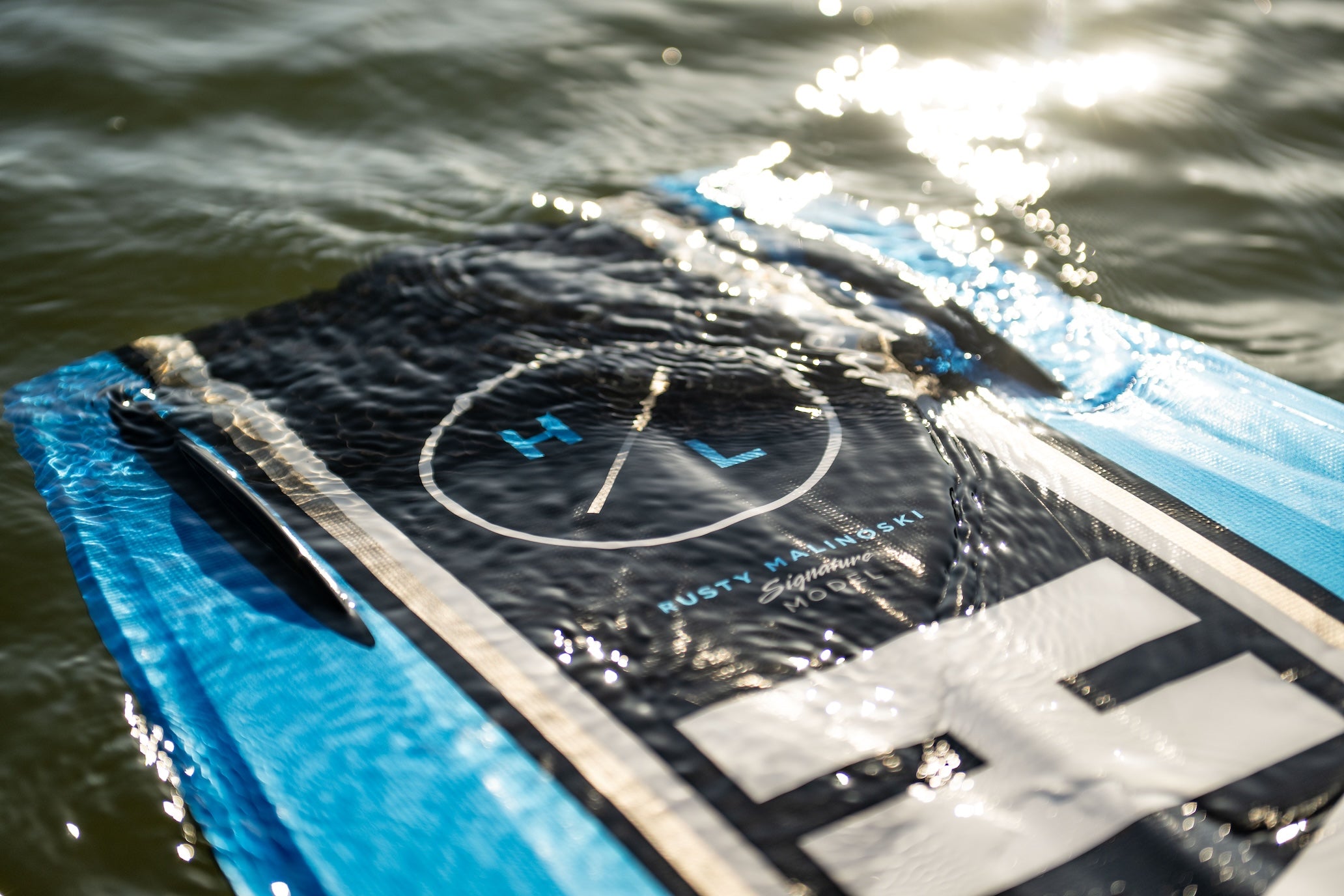 Close-up of wakeboard top floating in water with HL logo and Rusty Malinoski signature visible.