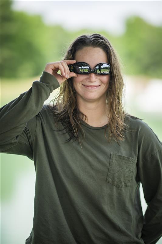 Person in a green shirt smiling and adjusting sunglasses outdoors.