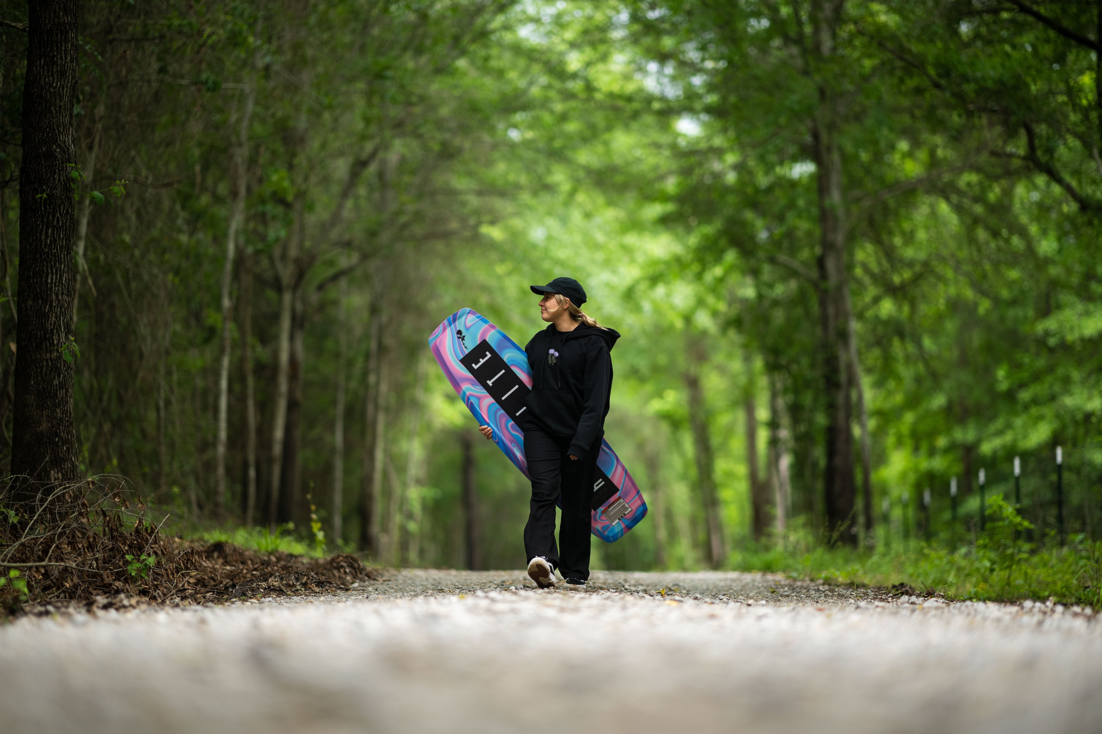 Person walking on forest path carrying a colorful wakeboard, wearing a cap and black clothes.