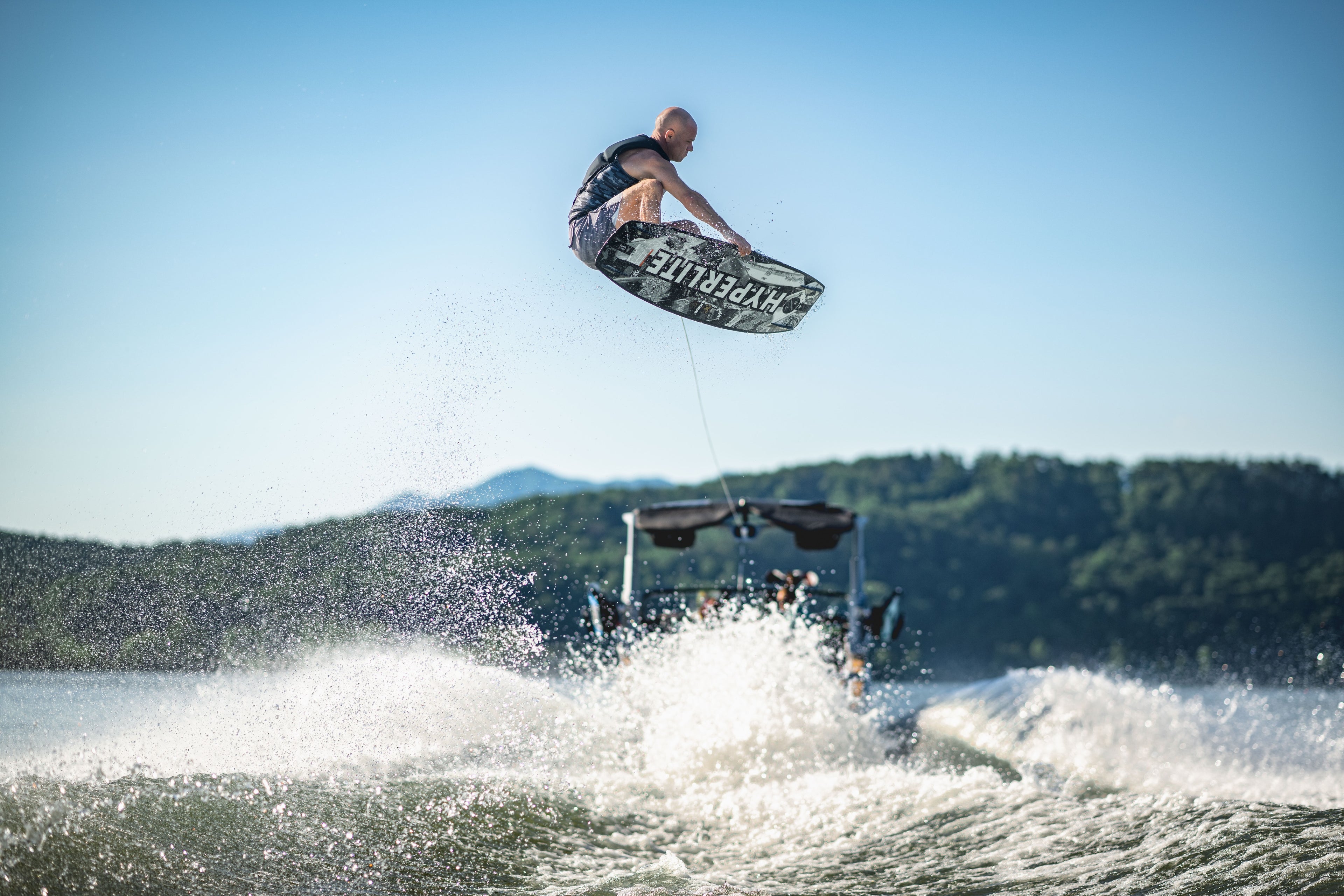 A person wakeboarding, airborne, with boat in the background, on a sunny day.