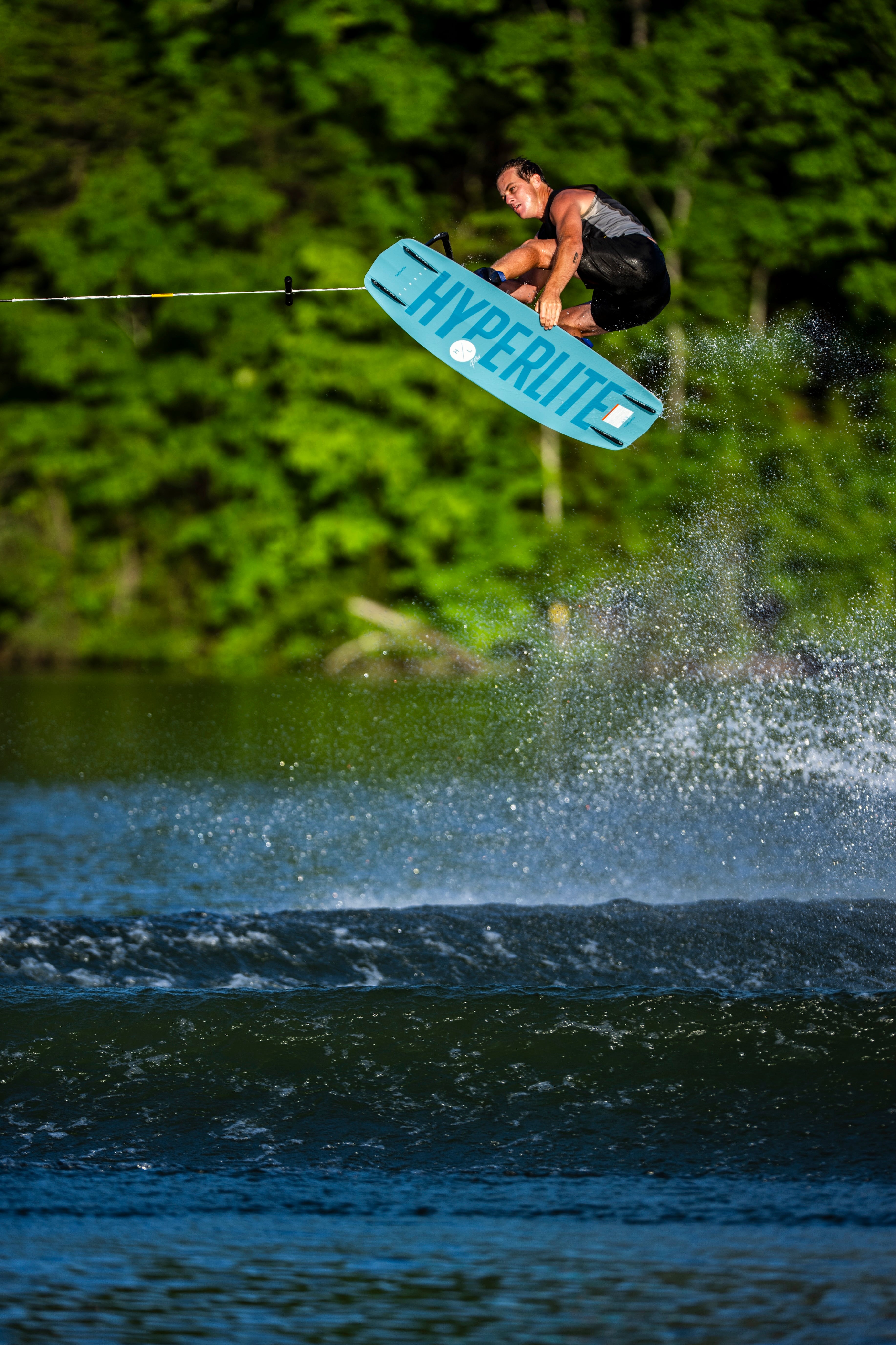 Person wakeboarding in the air over water, surrounded by lush green trees.
