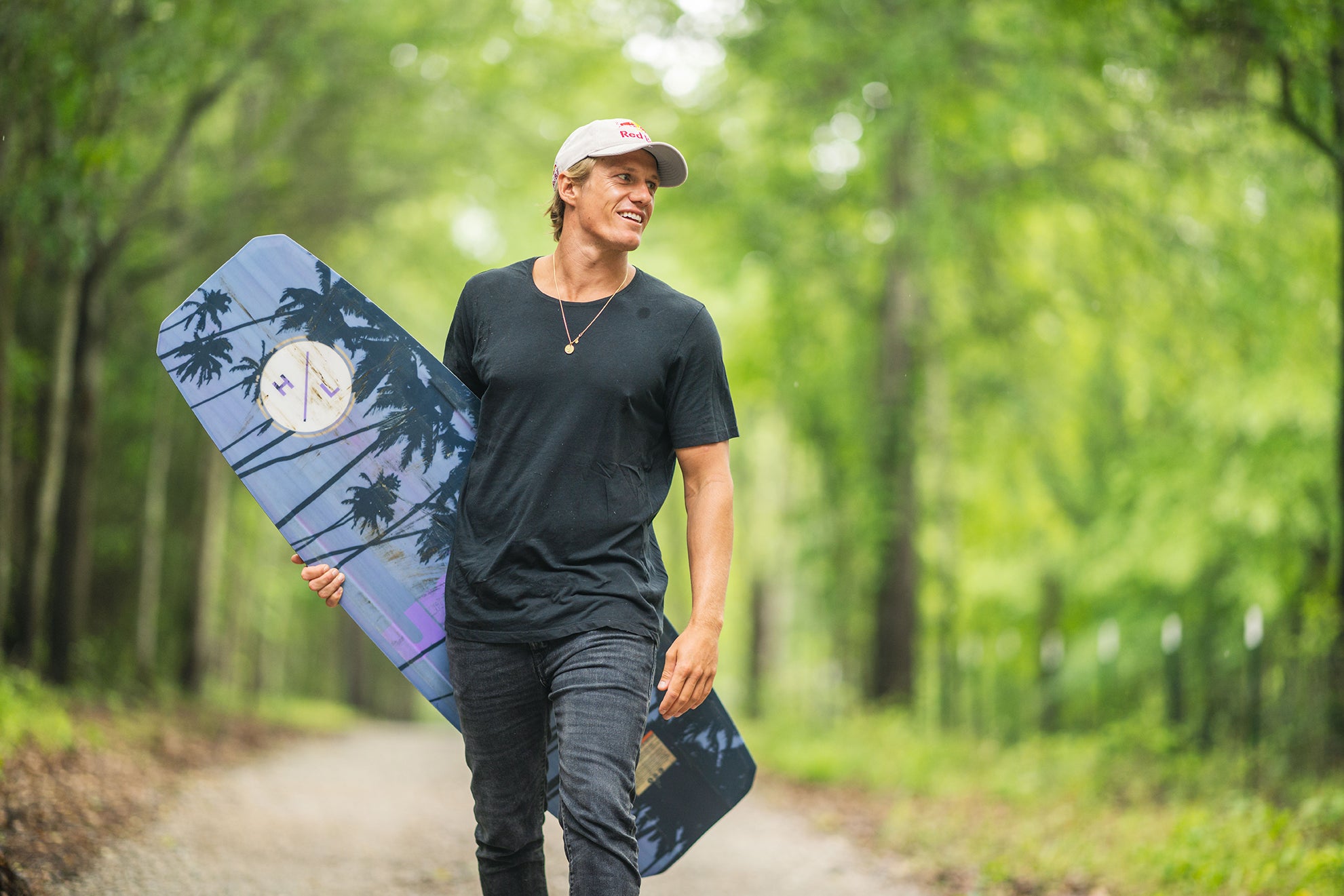 Person walking on a forest path holding a wakeboard, wearing a cap and black shirt.