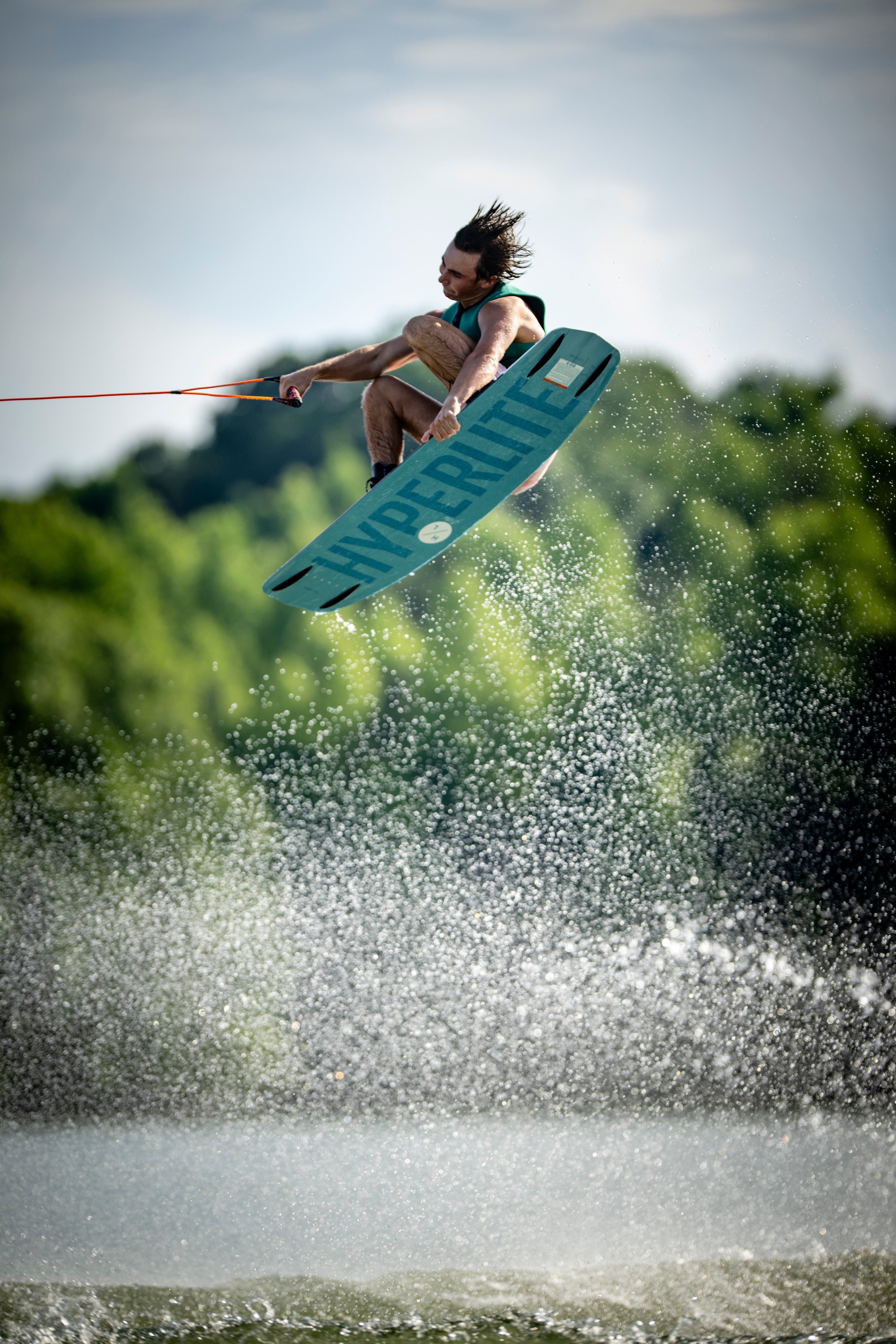Person wakeboarding mid-air with a bright sky and green trees in the background.