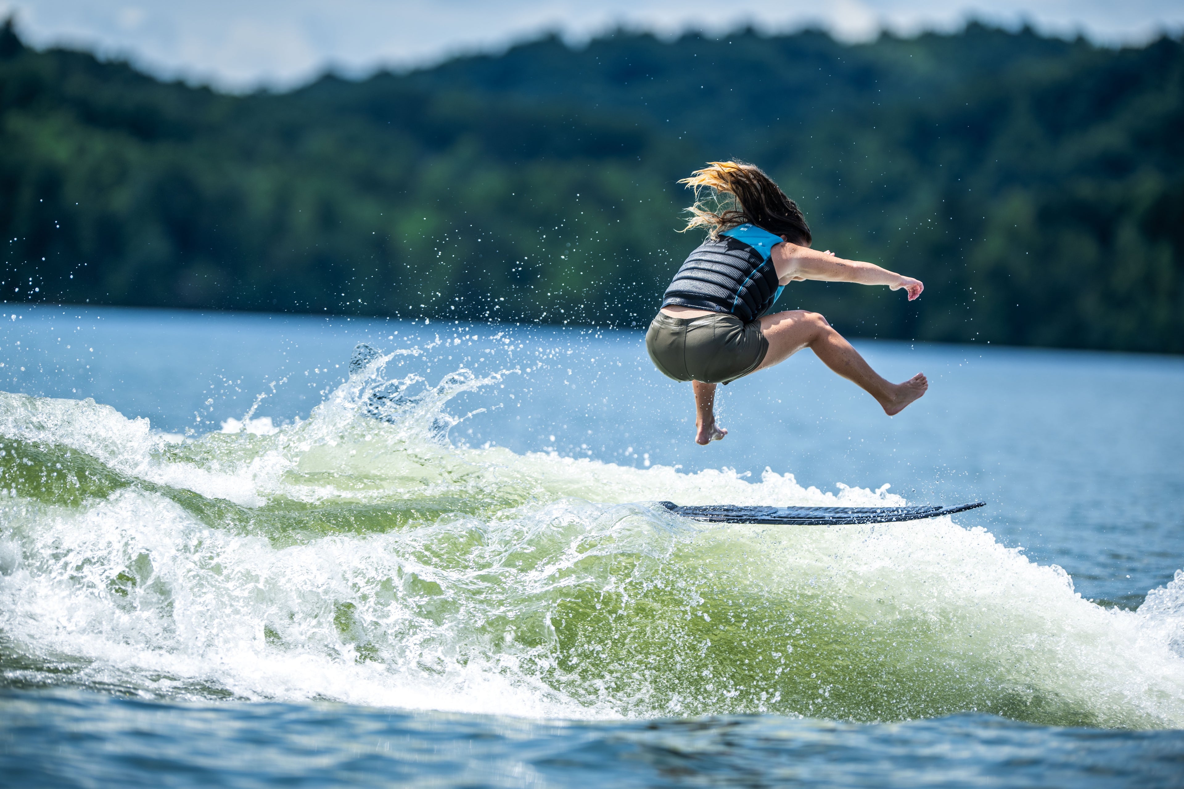 Person jumping off a surfboard on a lake with forested hills in the background.