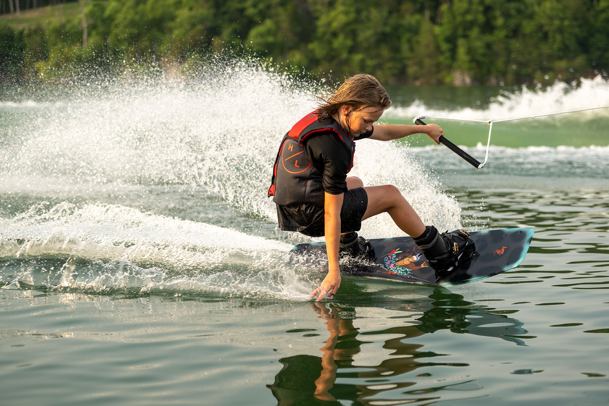 A person wakeboarding on a lake, creating splashes, surrounded by greenery.