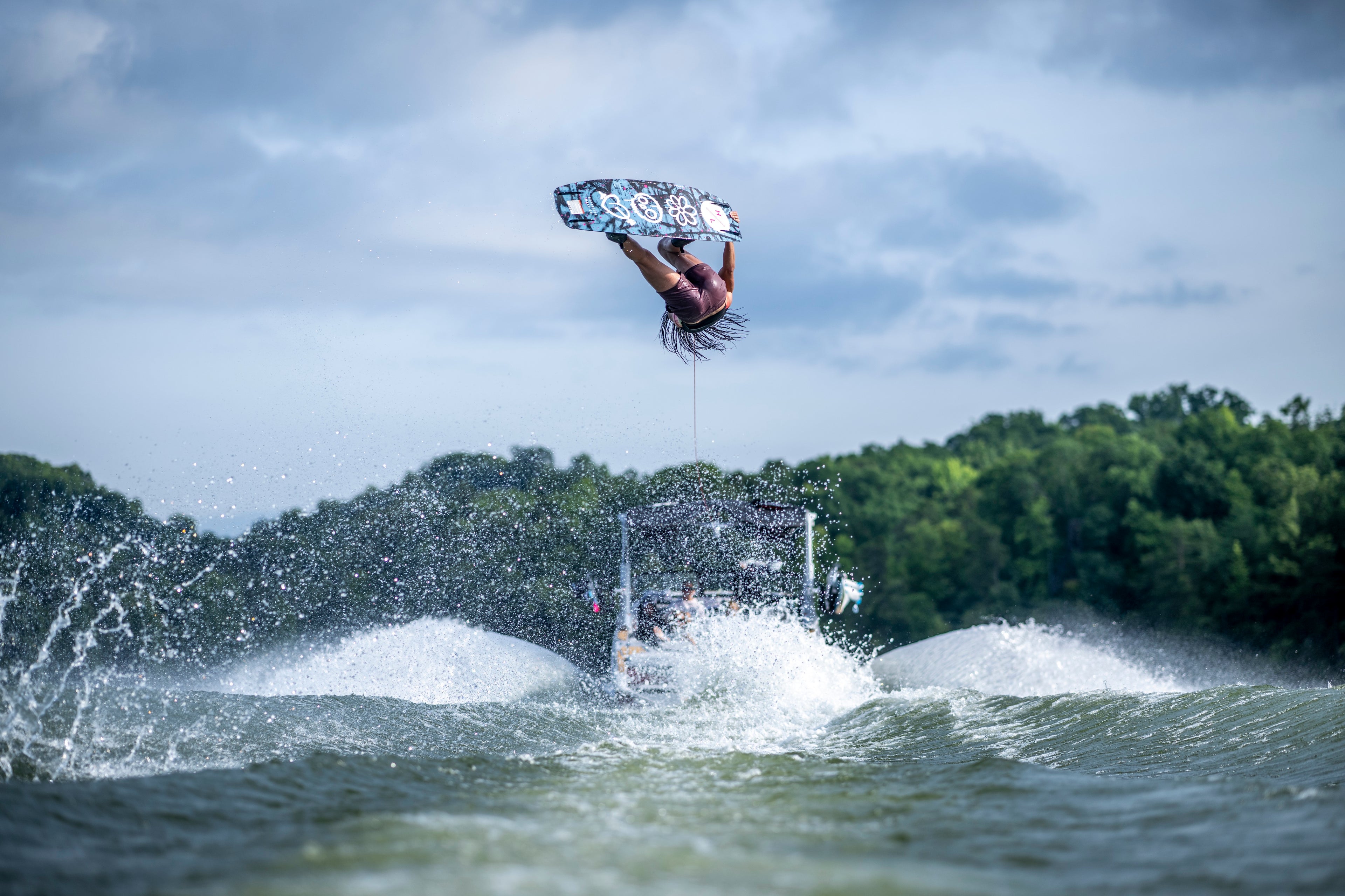 Person wakeboarding mid-air behind a boat on a lake, with trees and cloudy sky in the background.