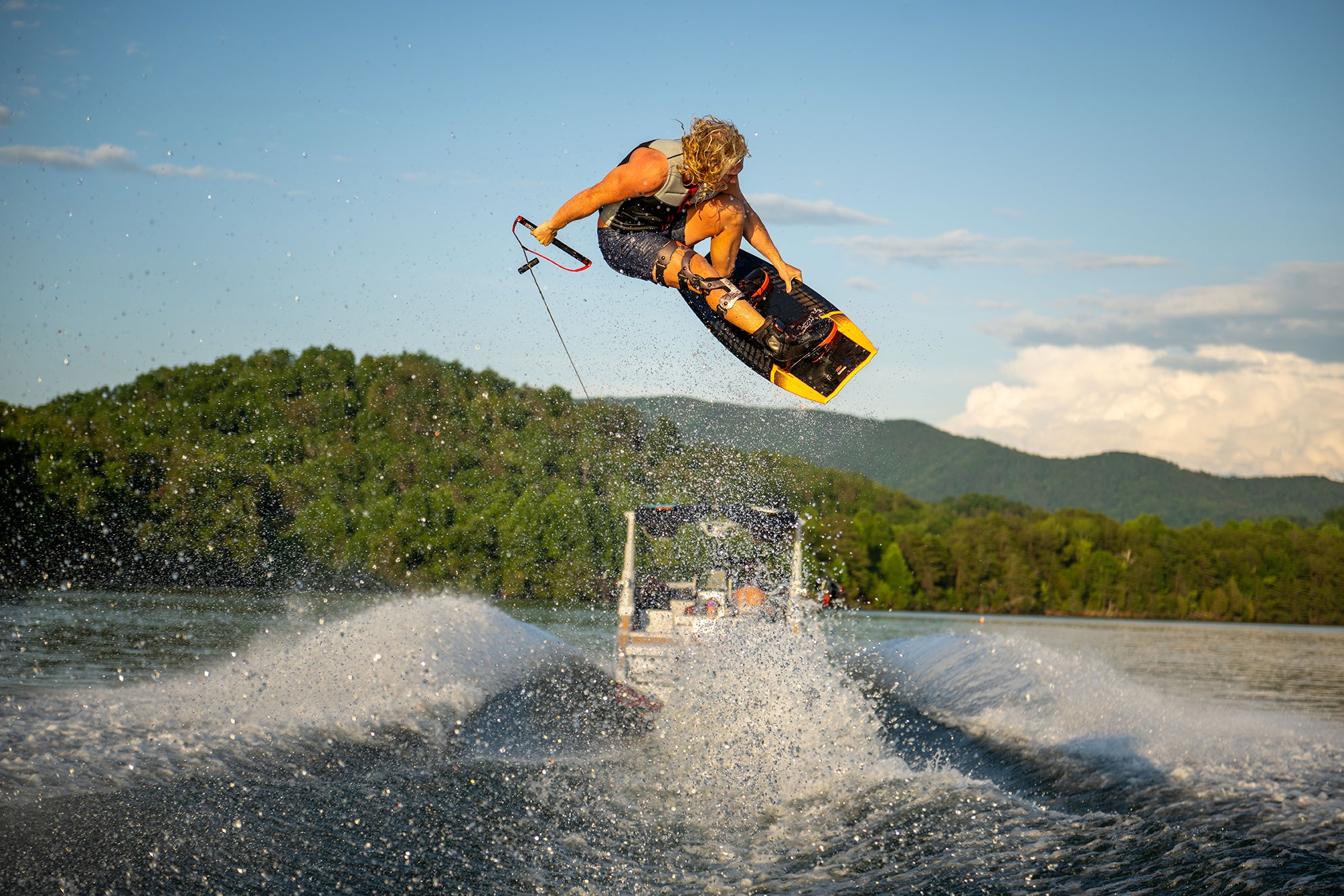 Person wakeboarding, mid-jump, with scenic lake and forested hills in the background.