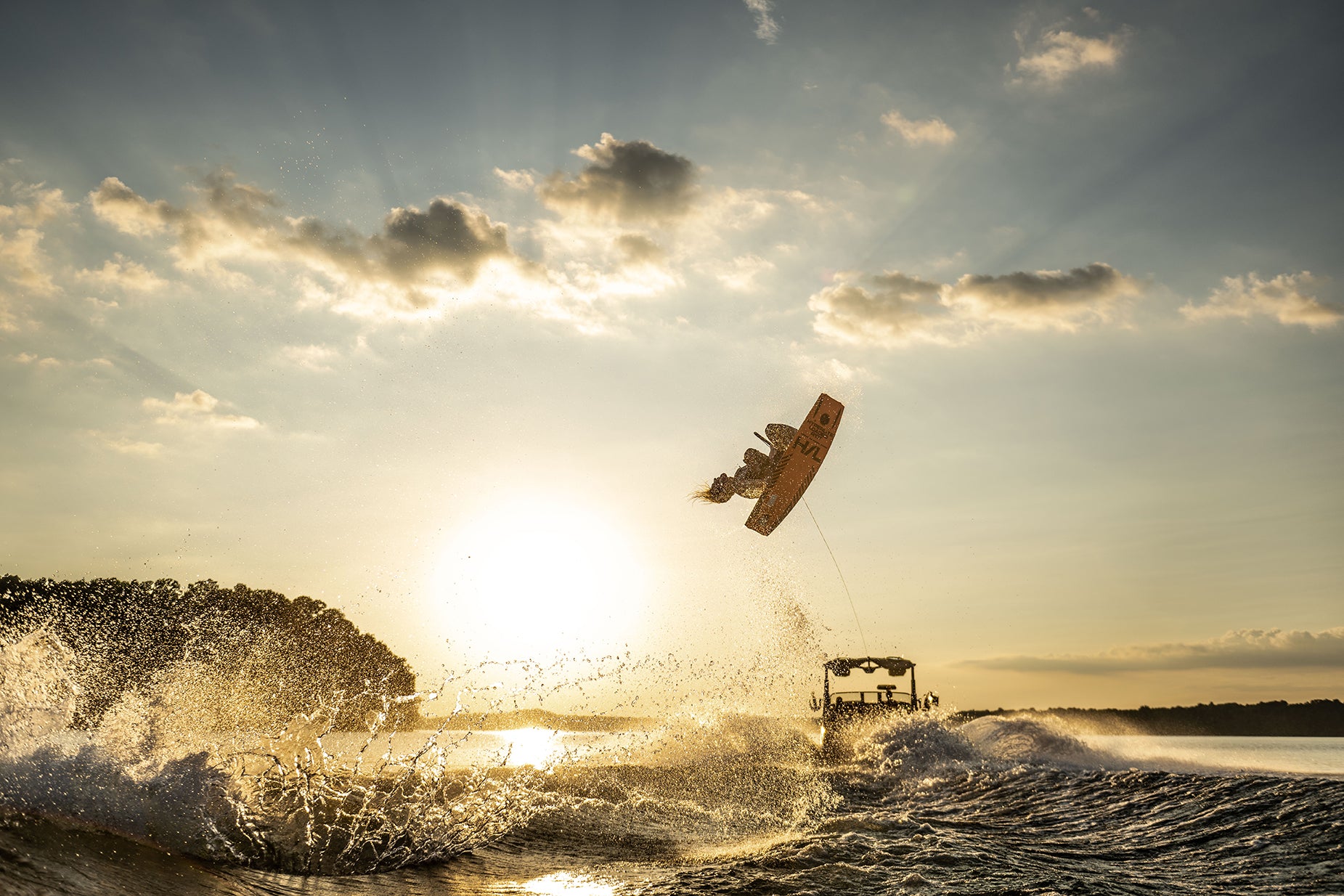 Person wakeboarding in the air at sunset, boat creating waves, silhouetted against the sun.