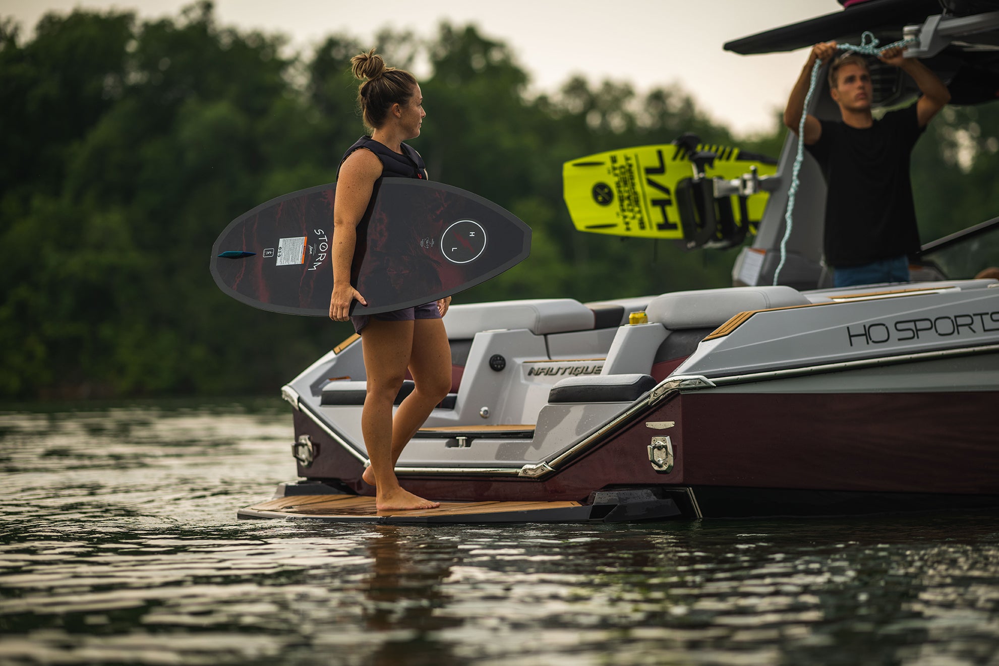 Person with a wakeboard on a boat at a lake, trees in the background.