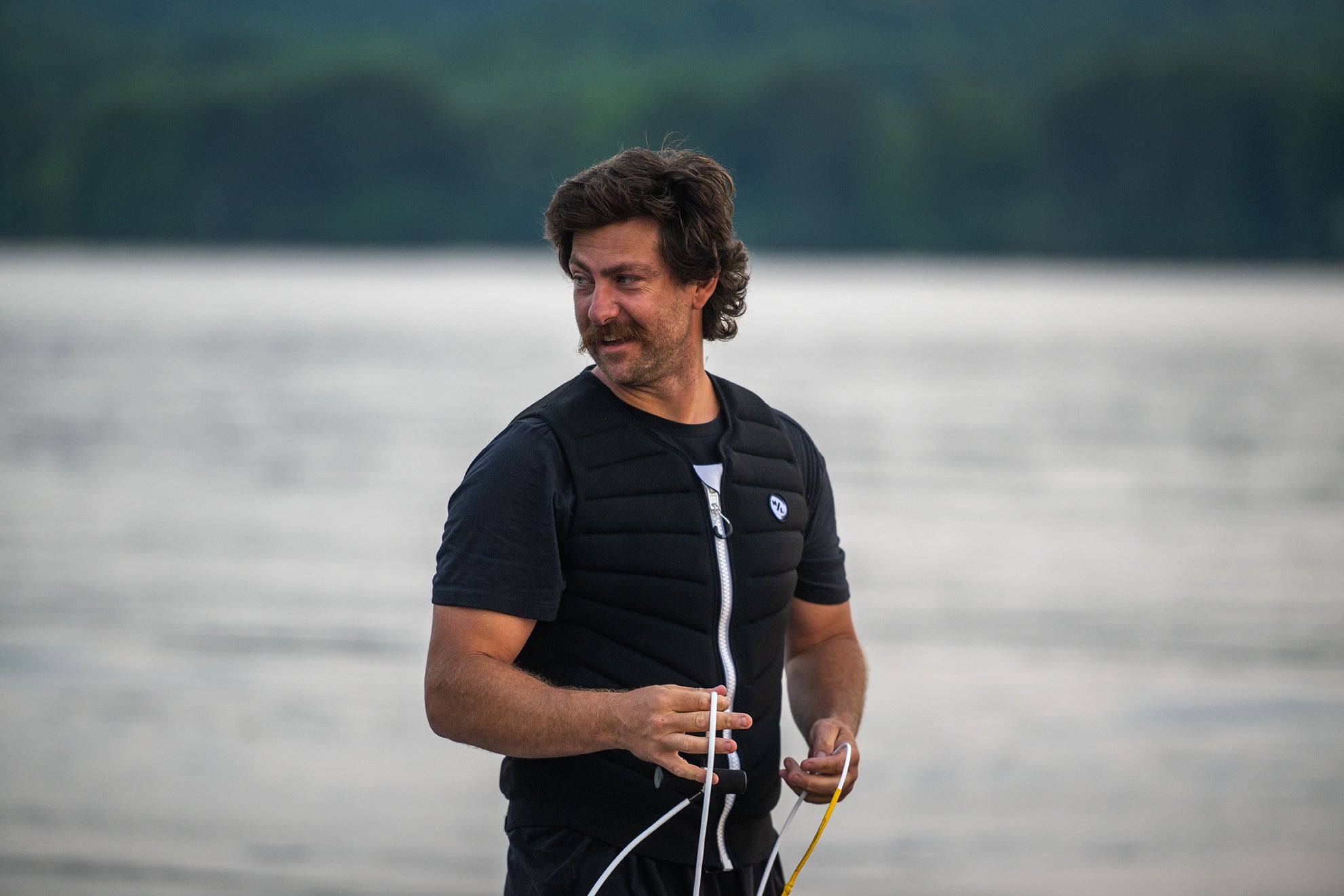 Man in a life vest holds a rope by a lake, with trees in the background.