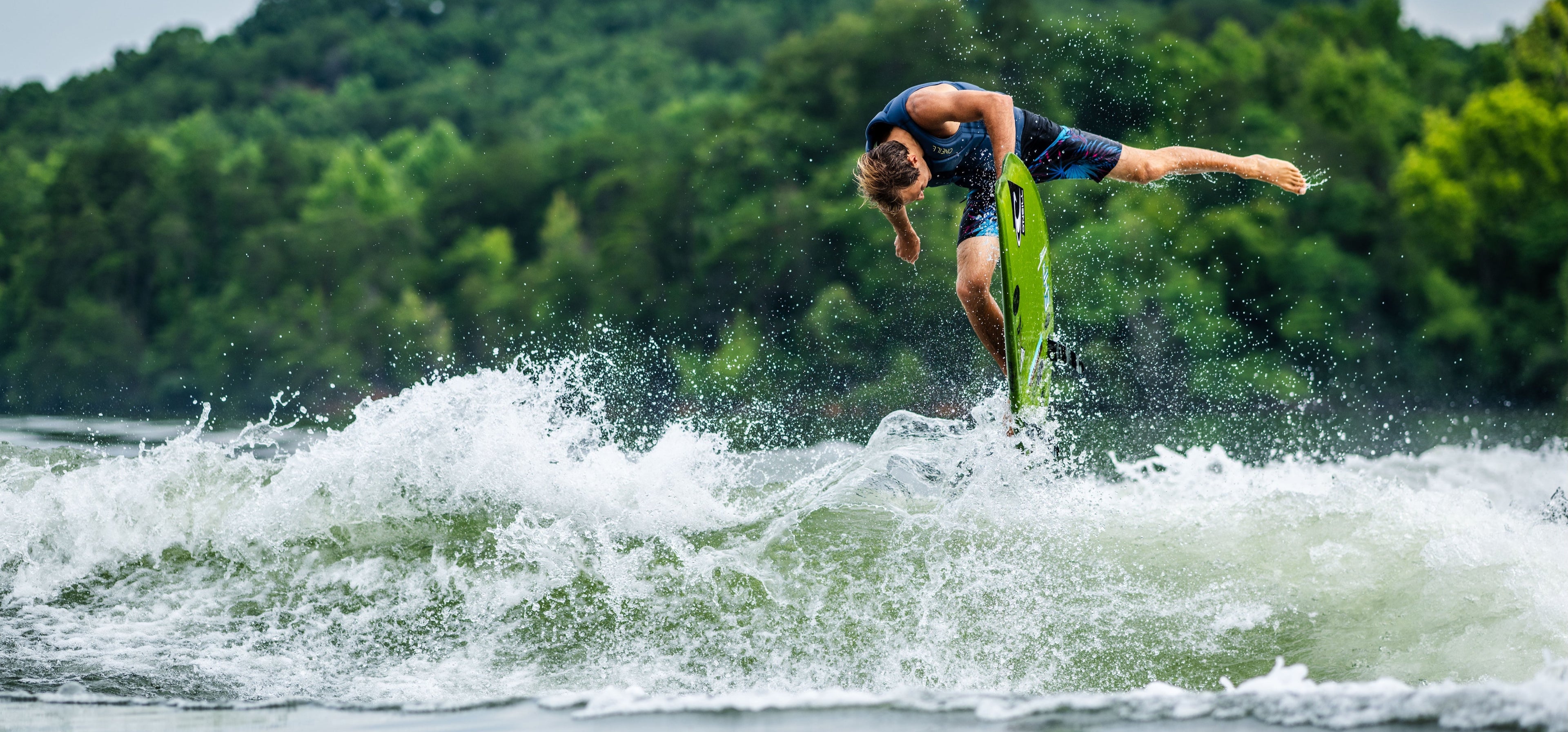 Person performing a trick on a surfboard amidst splashing waves and green foliage.