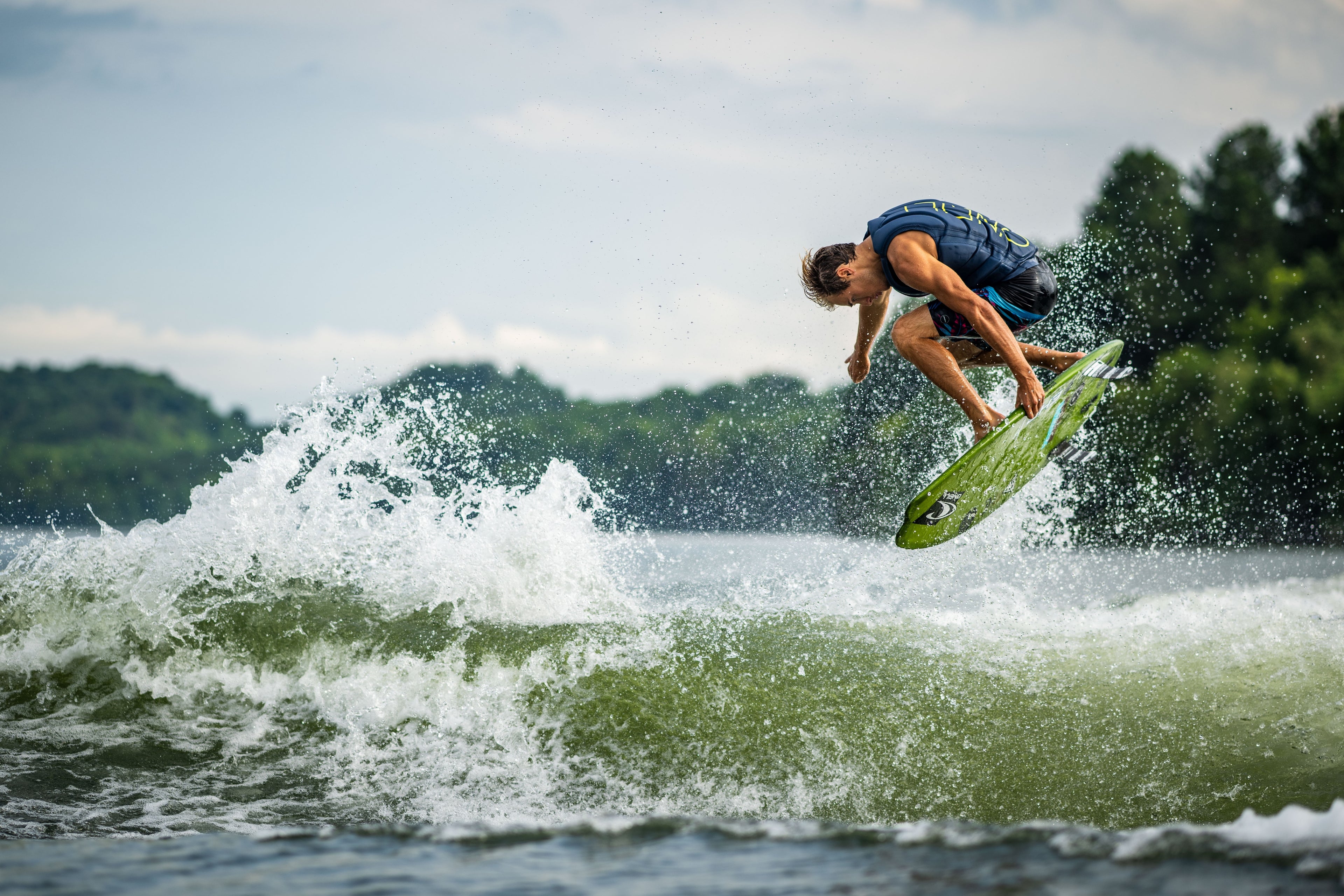 Person doing a mid-air trick on a surfboard over a wave near a forested shoreline.
