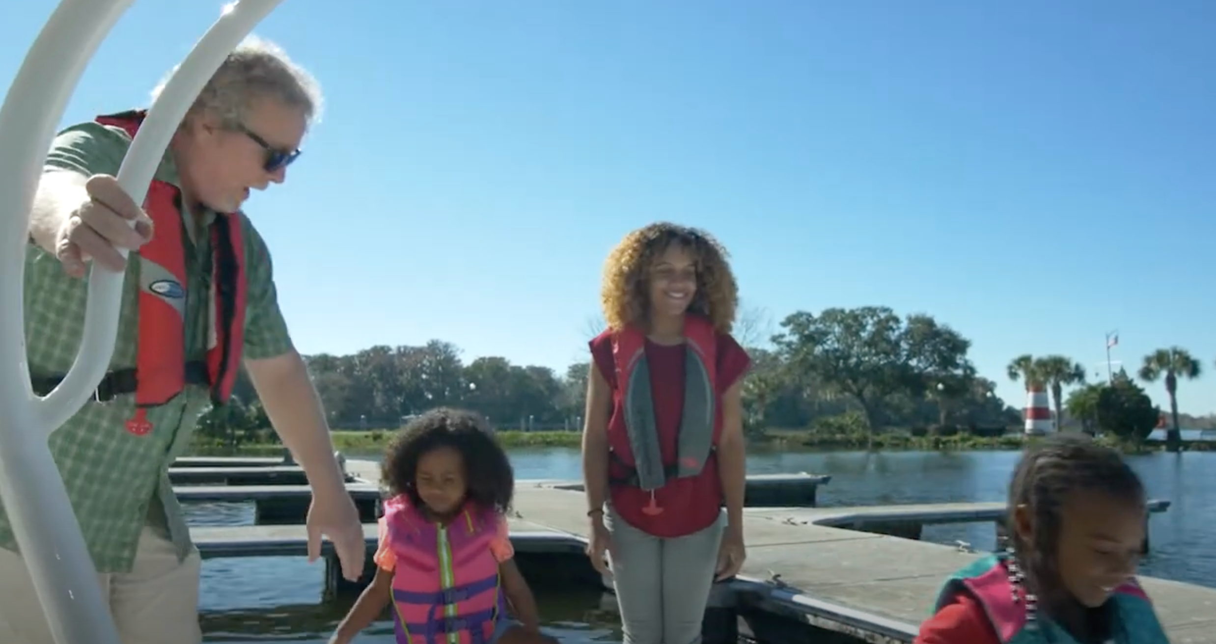 A family in life jackets stands on a dock near the water on a sunny day.