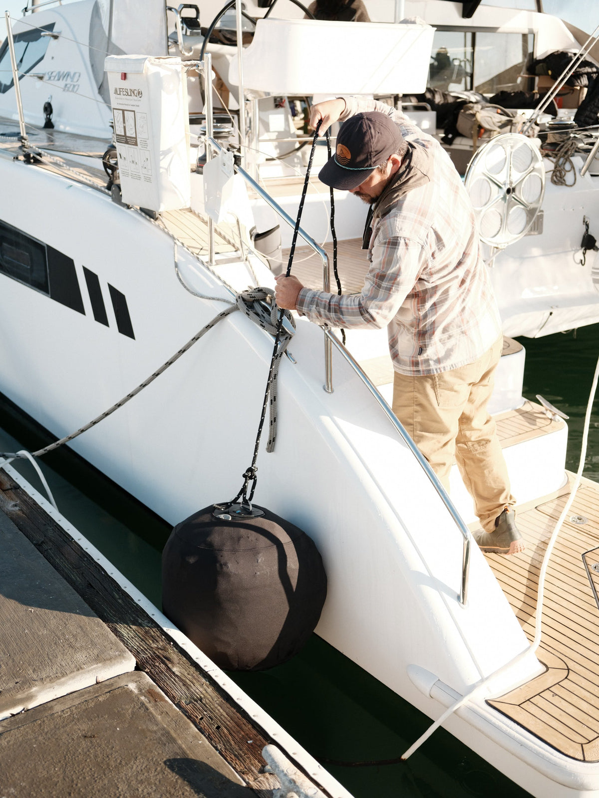 Person tying a black inflatable fender to a catamaran railing while docked at a marina.