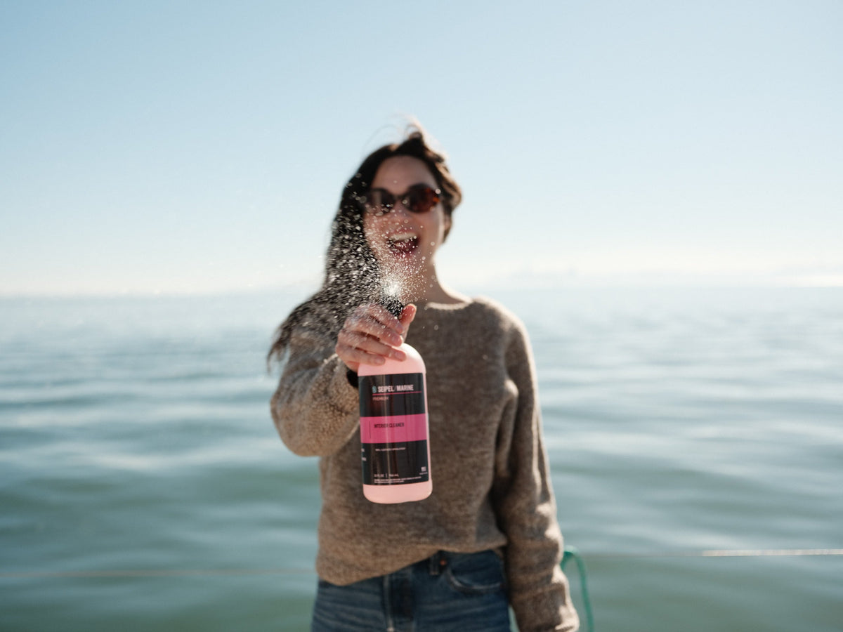 Person spraying marine cleaner toward the camera while standing on a boat with open water behind.