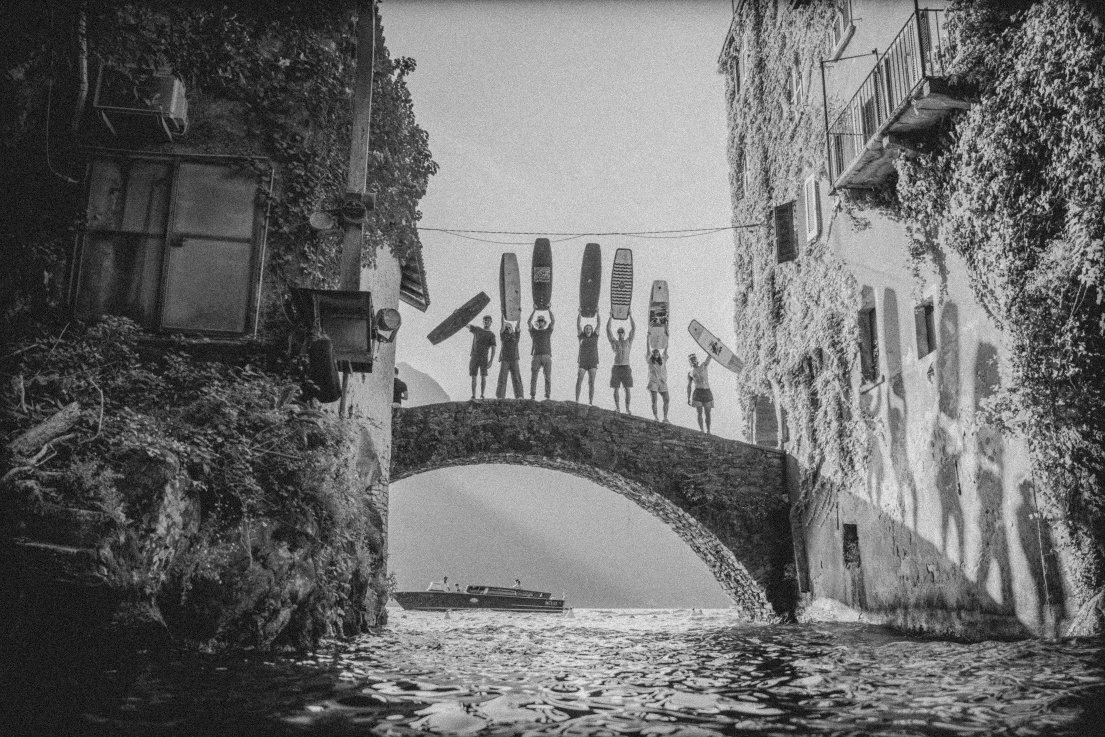 Black and white scene of a canal with a stone bridge with Hyperlite Pro riders holding their boards over their head with a boat in the background.