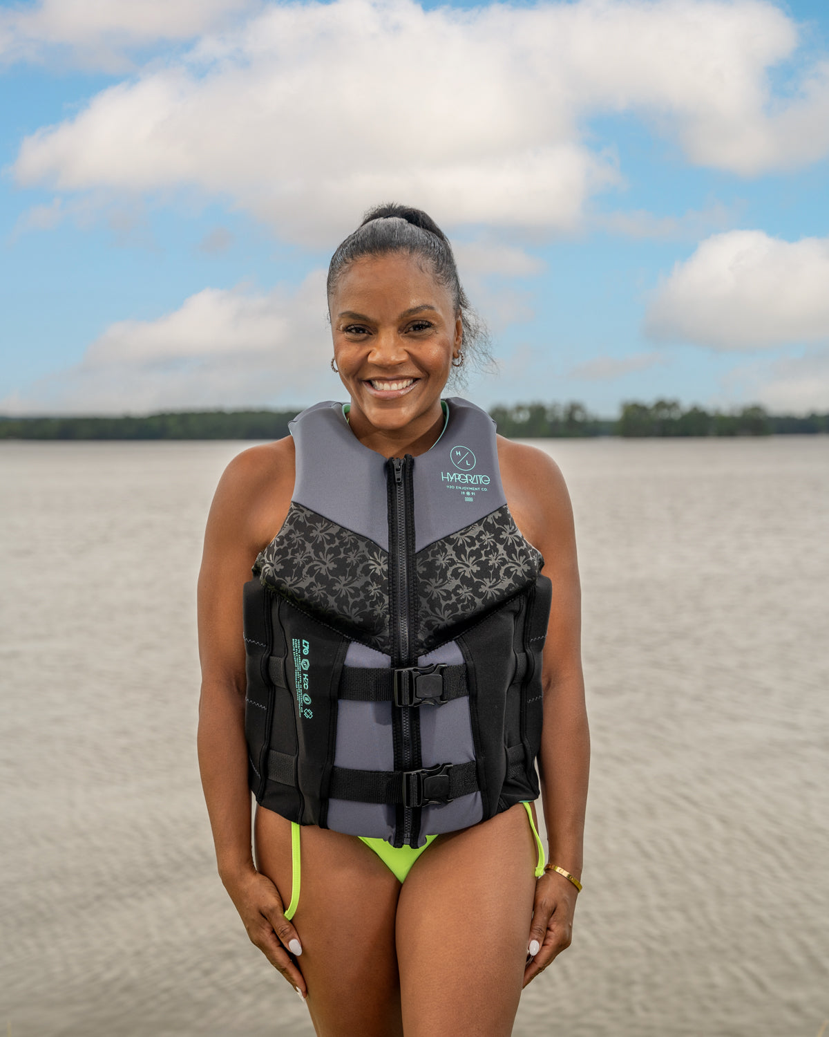 Smiling woman in a life jacket stands by the water under a partly cloudy sky.