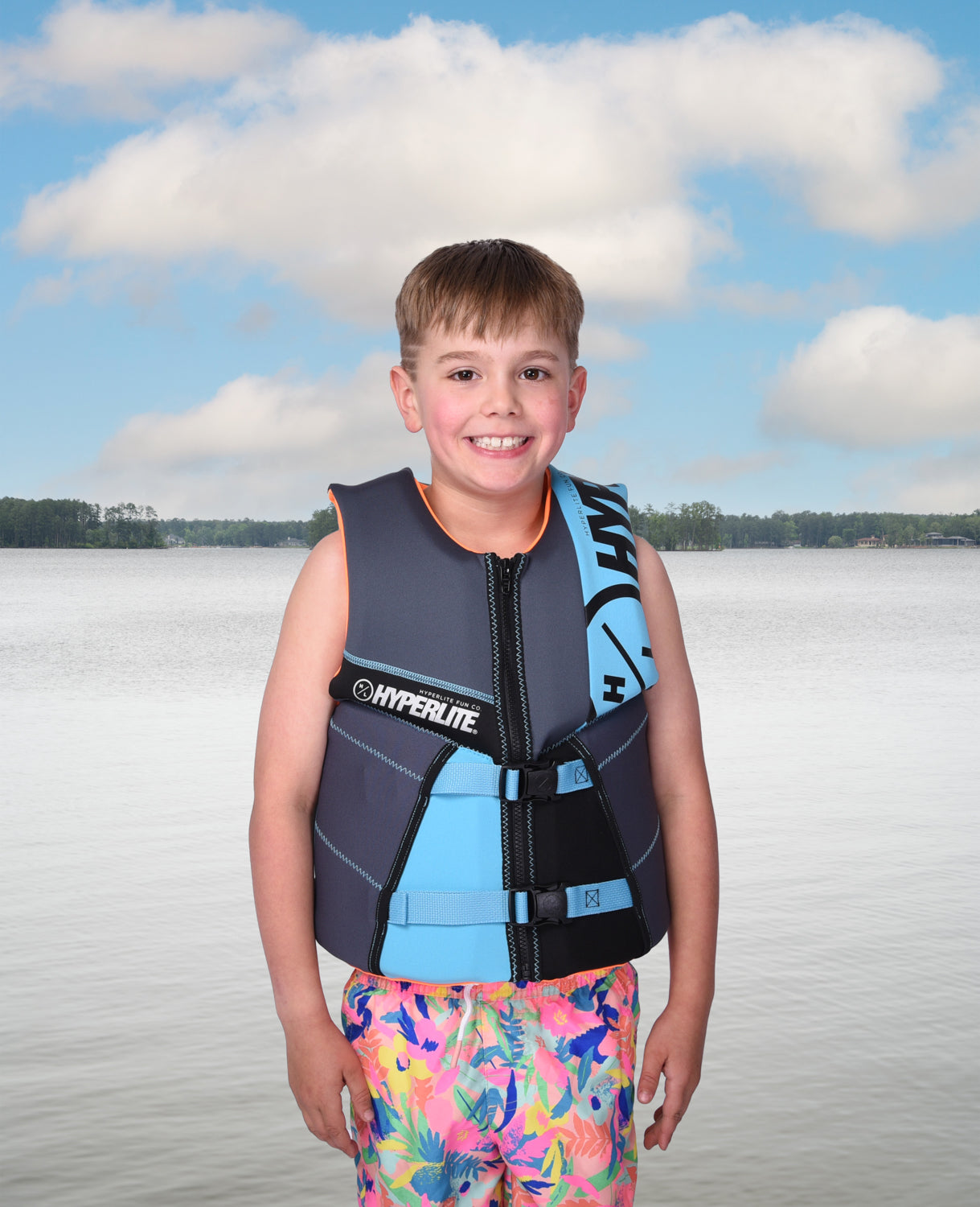 Boy in colorful shorts and life vest standing by a calm lake under a cloudy sky.