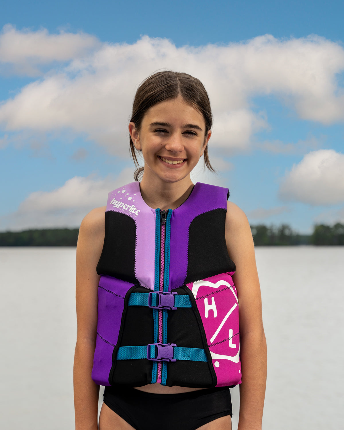 Child in a purple life jacket smiling by a lake with a cloudy sky.