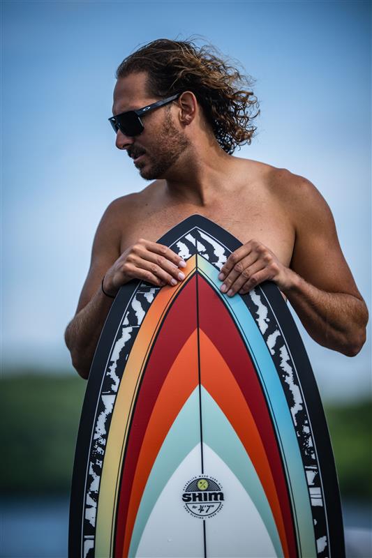 Shirtless man with sunglasses holding a colorful surfboard, against a blurred outdoor background.