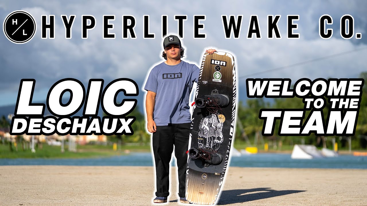 Person holding a wakeboard, standing on a sandy area with branding text in the background.